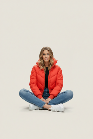 Red quilted puffer jacket with hood and front zipper; model seated with arms crossed.