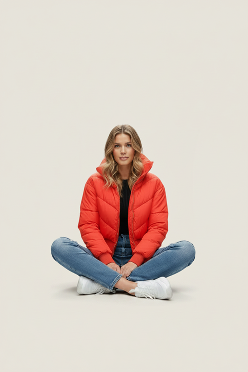 Red quilted puffer jacket with hood and front zipper; model seated with arms crossed.